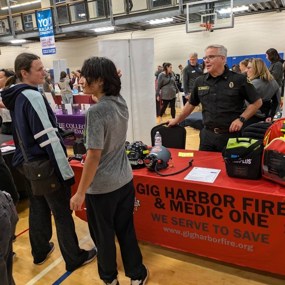 A Gig Harbor Fire & Medic One representative in uniform talks to a young student and an adult at a community event held in a gymnasium. The fire department's table displays helmets, equipment, and informational materials, with a red tablecloth that reads "Gig Harbor Fire & Medic One - We Serve to Save." People are gathered at other tables in the background, exploring various booths and interacting with presenters.