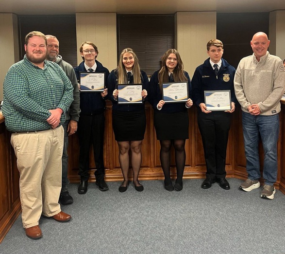 7 people stand in a row to pose for photo, with 4 holding framed certificates