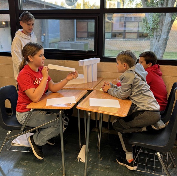 3 people seated at desks and 1 standing work on a paper tower