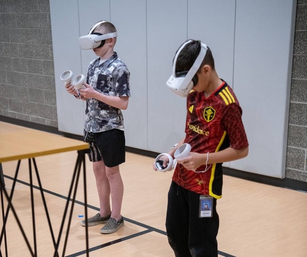 Two students wearing Meta Quest VR headsets stand in a gymnasium, each holding VR controllers. One student wears a gray camouflage shirt, while the other wears a red and yellow sports jersey. They appear to be participating in a virtual reality demonstration or activity.