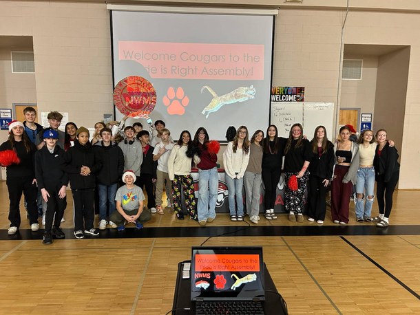 A group of students stand in a gymnasium, smiling for a photo. The banner behind them reads 'Welcome Cougars to the Pride is Right Assembly!'