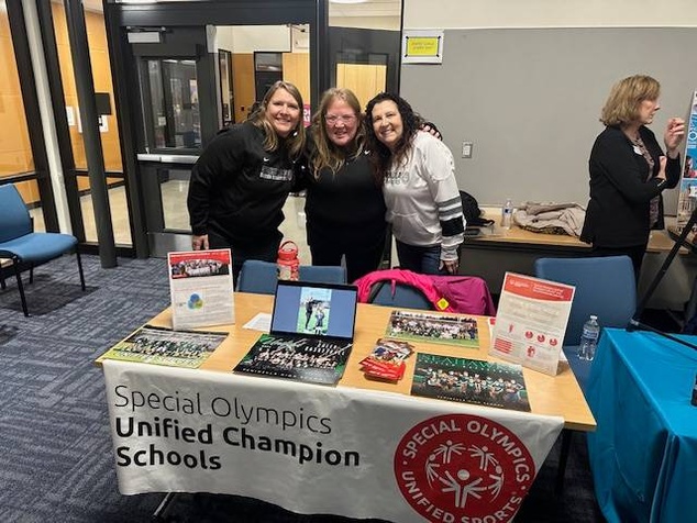 Three women lean together for a photo behind a table at a community event. The table has a laptop, flyers, and brochures on it with a banner that says "Special Olympics Unified Champion Schools"