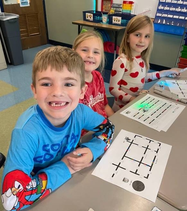 Three first grade students smiling, sitting at a desk with ozobots and color coding worksheet.