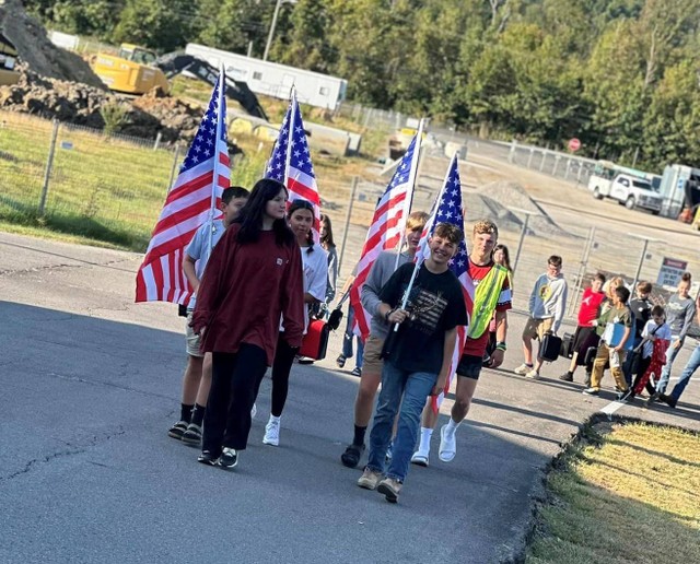 Group of about 16 people, with 4 carrying U.S. flags, walk up drive with construction in background