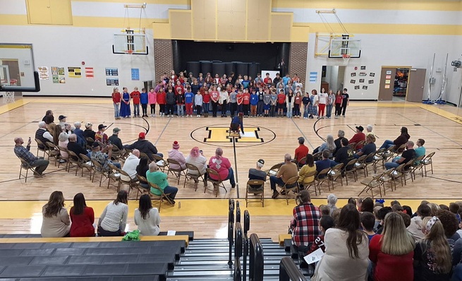 Students stand together in front of stage, with 3 rows of veterans sitting in front of them and others in the bleachers of the gym.