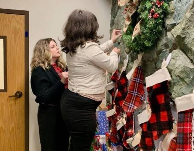 Two women decorate a wall with a wreath and stockings for the holidays.