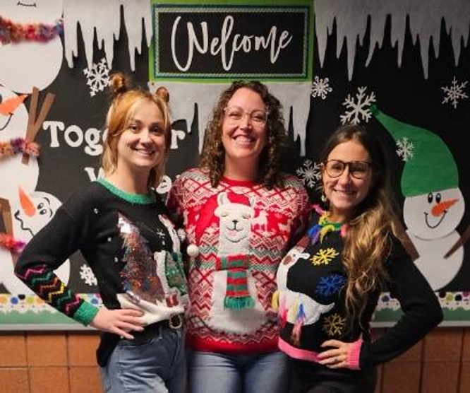 Three women wearing festive sweaters stand in front of a winter-themed bulletin board that says 'Welcome'.