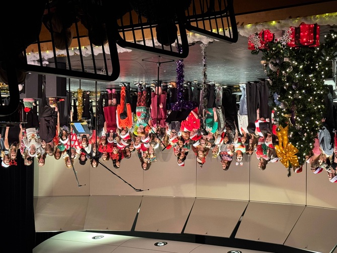 A large group of people, many wearing festive hats, stand in front of a decorated Christmas tree on a stage.