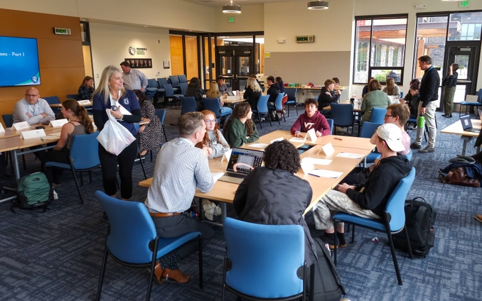 A large group of students and district officials sit in a conference room around multiple tables with computers and paper in front of them. They are in active discussion. Supt. Krestin Bahr is walking around the tables with a white grocery bag passing out candy.