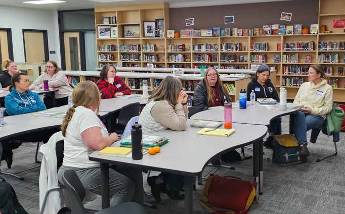 A group of adults sit at tables in a school library, with notes and water bottles on the table, in a discussion.