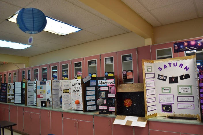 A classroom display of science fair projects about planets, with a blue paper lantern hanging from the ceiling.