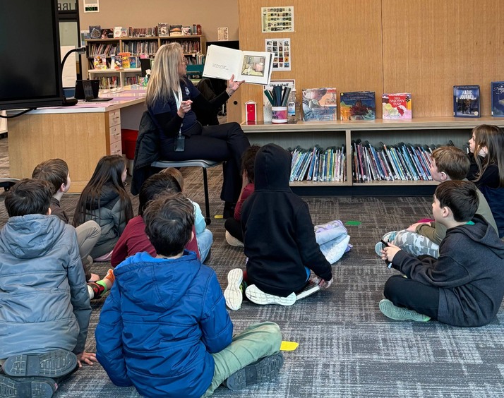 A group of children listen attentively as a Superintendent Bahr reads a book in a library setting.