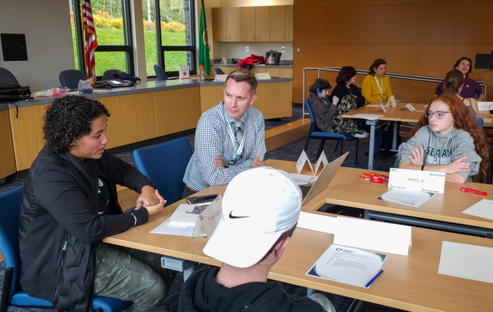 A student actively speaks with hand gestures to a table with two other students and a district official. There is paper and computers on the table, and other tables of district employees and students at tables around them in a large meeting room.