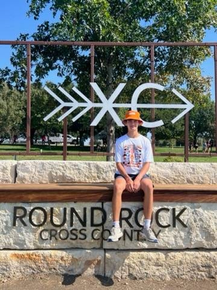 A young man sitting for a picture in front of the Round Rock Cross Country sign.