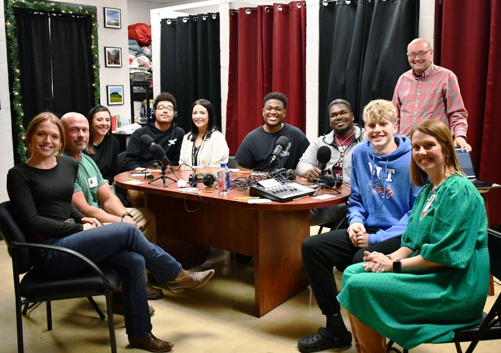 10 people sit at table with recording equipment.