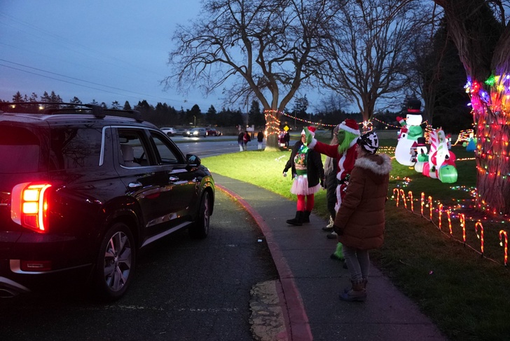 A car drives past people dressed in festive costumes and a tree decorated with Christmas lights.