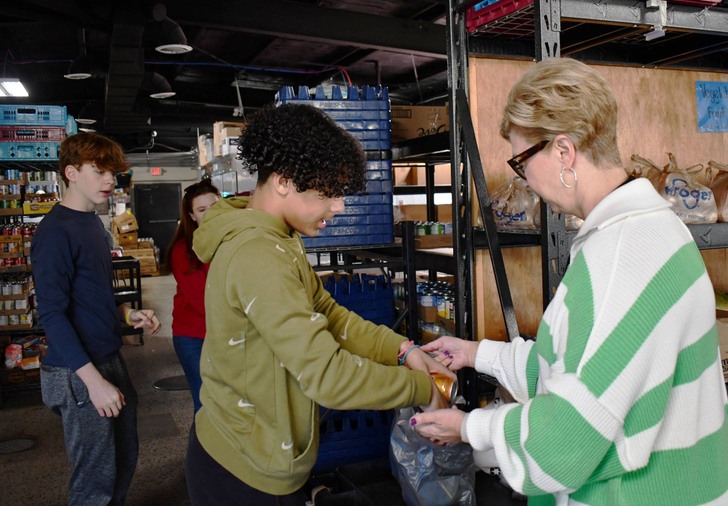 One person holds plastic bag while another puts cans in it. Two others stand in background.