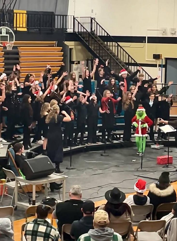 A choir performs on stage with a Grinch character in a festive setting.