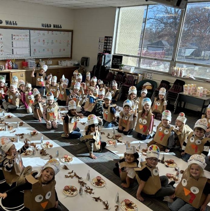 Dozens of kindergartens sit at floor tables together, with paper plates full of snacks for each student, and they're all wearing paper bag and construction paper turkey costumes.