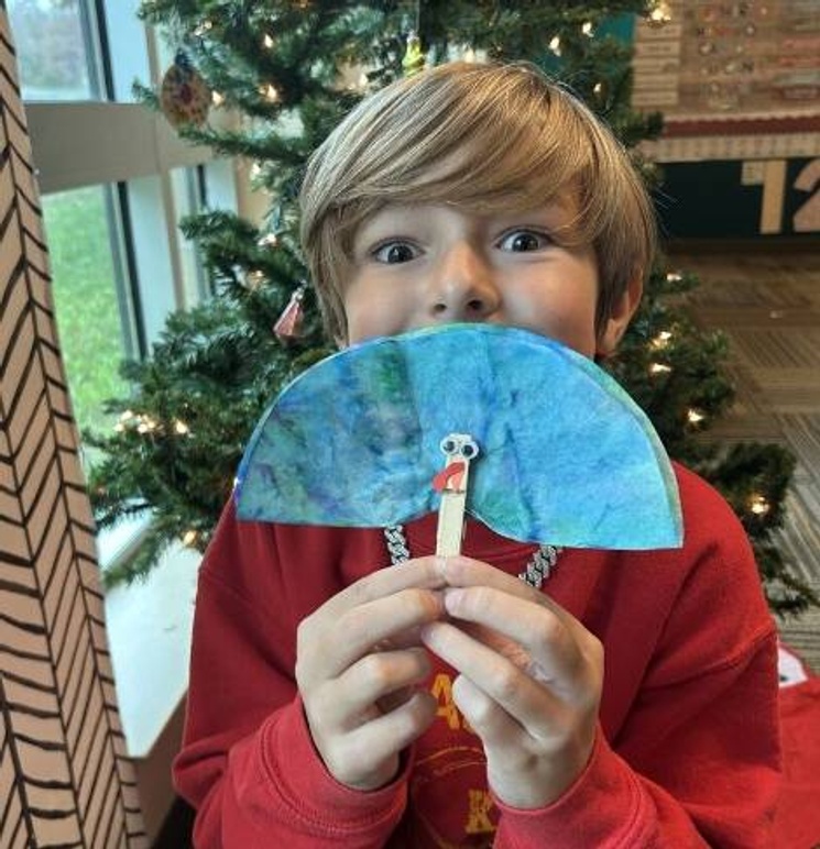 A young boy is holding a handmade clothespin turkey in front of his face, only his smiling eyes show behind the turkey's blue feathers.