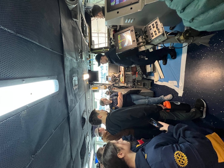 Inside the USS Nimitz, several students tour the space, with one wearing a Rotary Club sweatshirt. They are listening to crew members explain the ship's navigation equipment and controls. Electronic displays and control panels are visible throughout the space, and the ceiling shows typical maritime vessel infrastructure with lighting and ventilation systems.