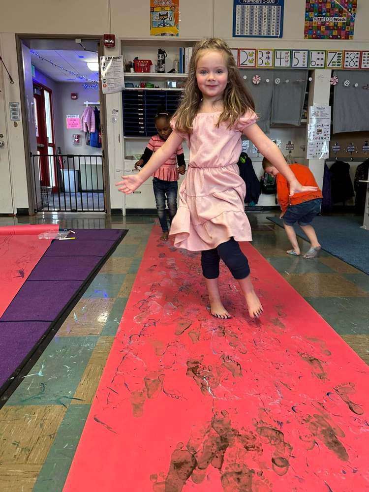 A young girl walks barefoot on a red paper path covered in footprints, with other children in the background.