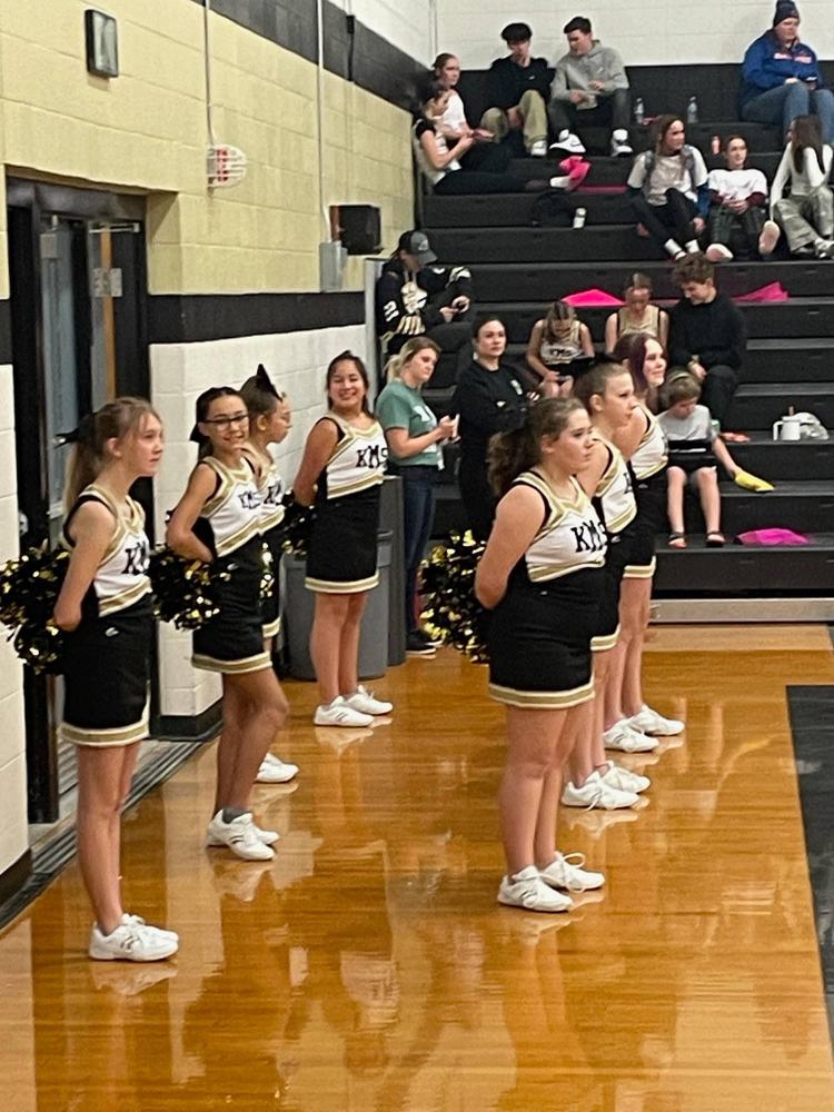 A group of cheerleaders in black and gold uniforms stand in a line on a basketball court, holding pom-poms.