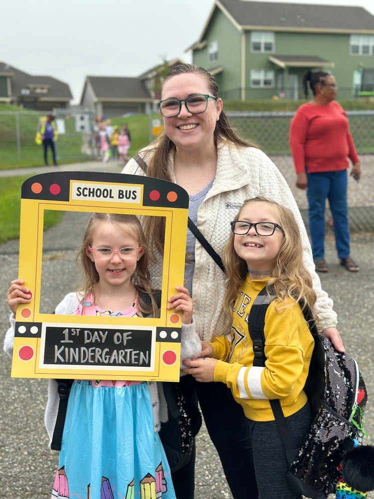 mother and two young daughter pose with first day of kindergarten sign that looks like a school bus with cutout in the center