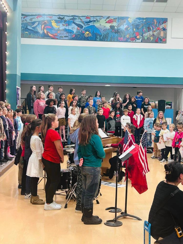 A large group of elementary students stand in a gymnasium on choir stands with U.S. Flags, a drum set and a piano singing during an event.