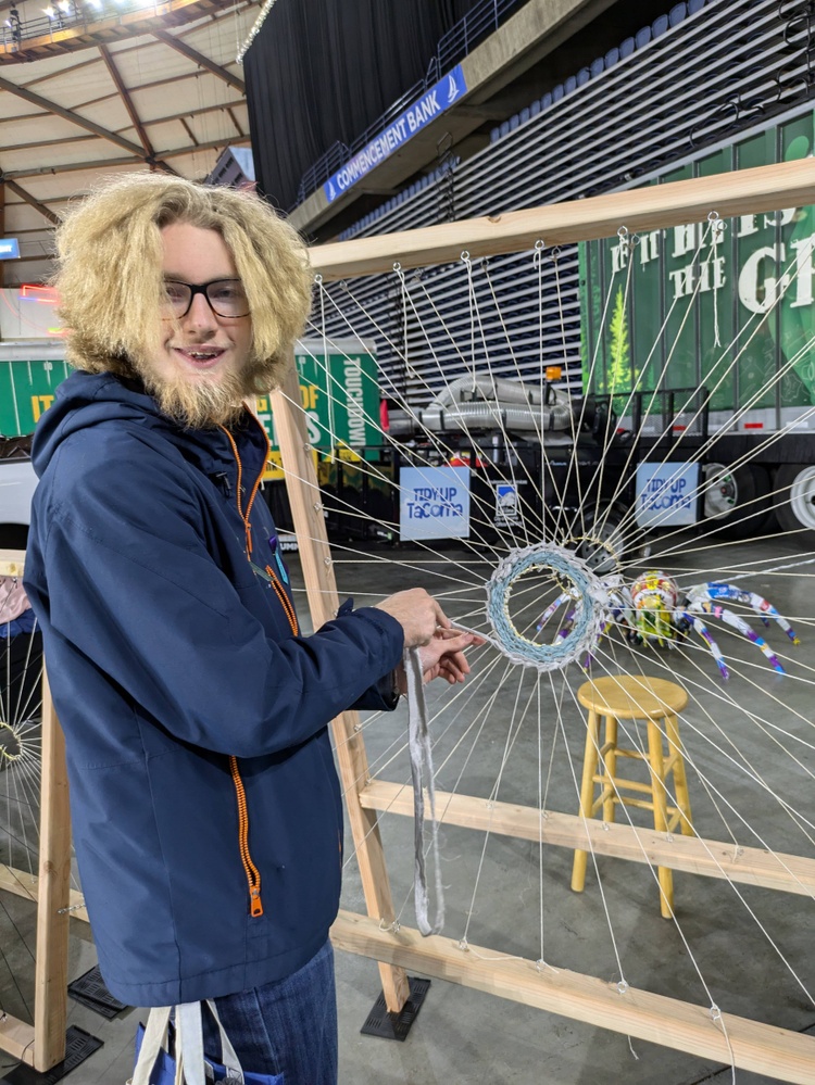 A high school student with long blond hair, a blond beard, and wearing glasses smiles for a photo as they interact with a weaving project during a career fair in a large arena.