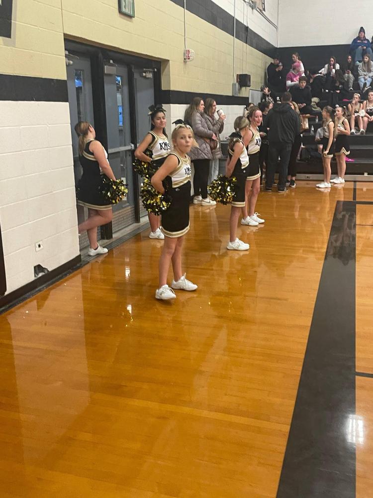 A group of cheerleaders in black and gold uniforms stand on a basketball court, holding pom-poms.