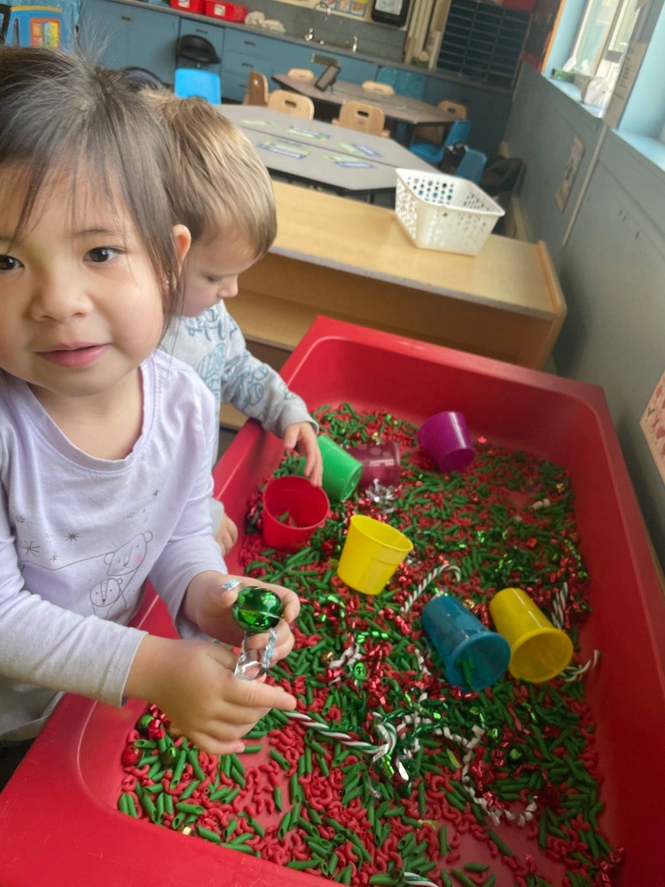 Two young children play in a sensory bin filled with green and red pasta, colorful cups, and festive decorations.