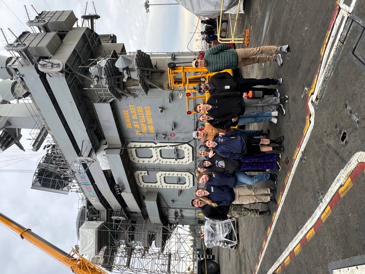 A group of students and adults pose for a photo on the deck of the USS Nimitz. They stand in front of the ship's tower structure, which has visible warning text reading 'BEWARE OF JET BLAST PROPELLERS AND ROTORS' and construction scaffolding. A yellow crane and various equipment are visible in the background. The group appears to be on a tour of the vessel, with safety lines and deck markings visible in the foreground.