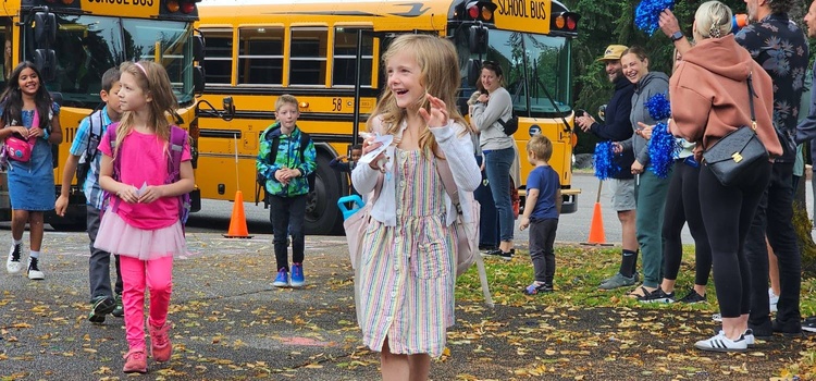 Young students stepping off school buses, greeted with excitement by cheering parents and staff holding blue pom-poms. The joyful atmosphere captures the start of the school day, with smiling children carrying backpacks, ready for a day of learning.