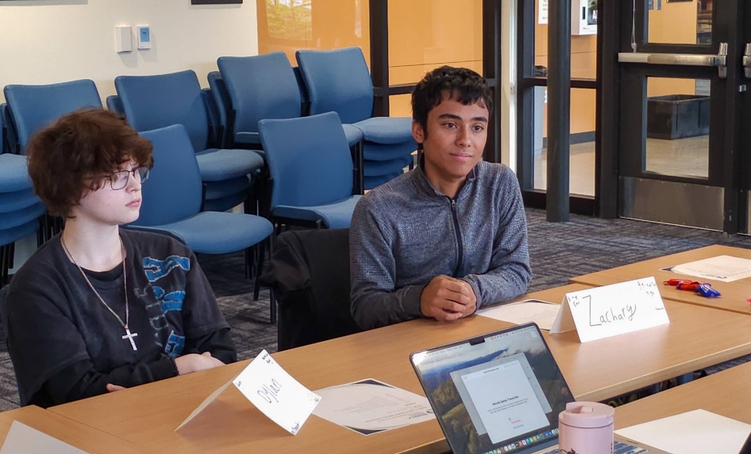 Two high school students sit next to each other at a table in a conference room, with papers and markers on the table. They are speaking with someone out of frame who has a laptop in front of them. 