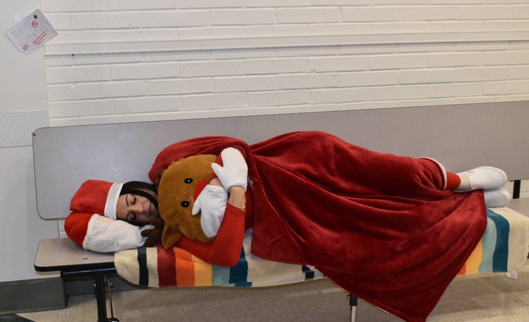 A person wearing a Santa hat and red blanket sleeps on a bench with a reindeer plush toy.