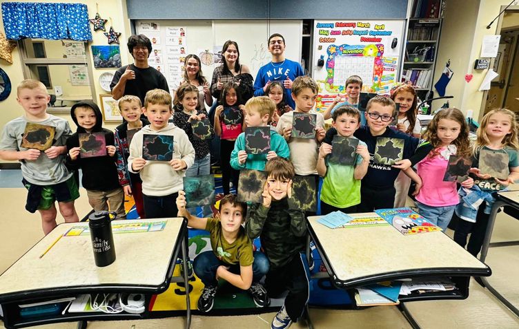 A large group of students stand together holding up a rainbow leaf art project. High school students stand in the back and second grade students stand in the front. The photo was taken inside an elementary classroom.