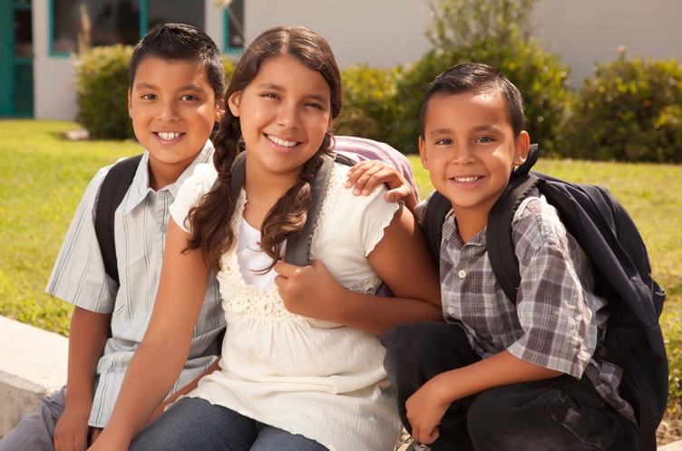 Three children with backpacks smile for the camera outside a school building.