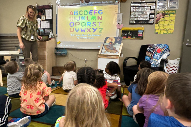 A class of preschoolers sits on a carpet before a smartboard showing playfully-designed ABC's, with a teacher beside it, interacting with the students.