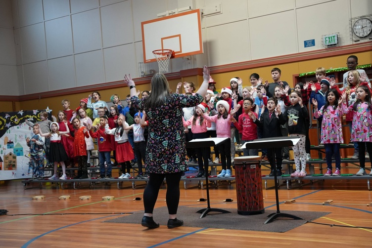 A group of children sing with their teacher in a gymnasium, with a basketball hoop in the background.