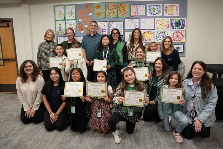 A group of children and adults pose for a photo, holding certificates of recognition. The children are smiling and looking at the camera.