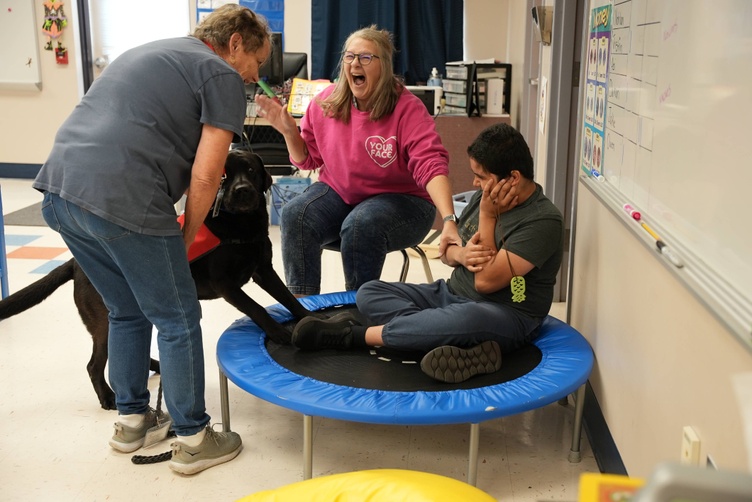 staff member laughing out loud and student smiling while being greeted by a black dog