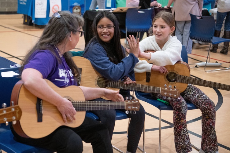 Three people sit in blue chairs in a school gymnasium, with two acoustic guitars. An instructor in a purple shirt is showing something on the guitar to two students who are smiling and engaged in the musical instruction.