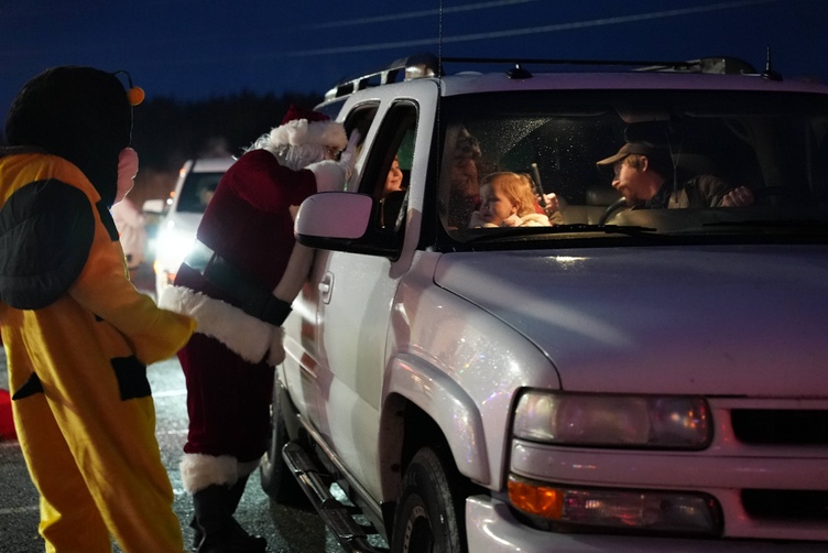 Santa Claus greets a child in a car at a drive-thru event, with a person dressed as a bee standing nearby.