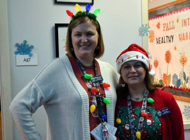 Two women wearing festive holiday attire smile for a photo. One woman wears a headband with lights and the other wears a Santa hat.