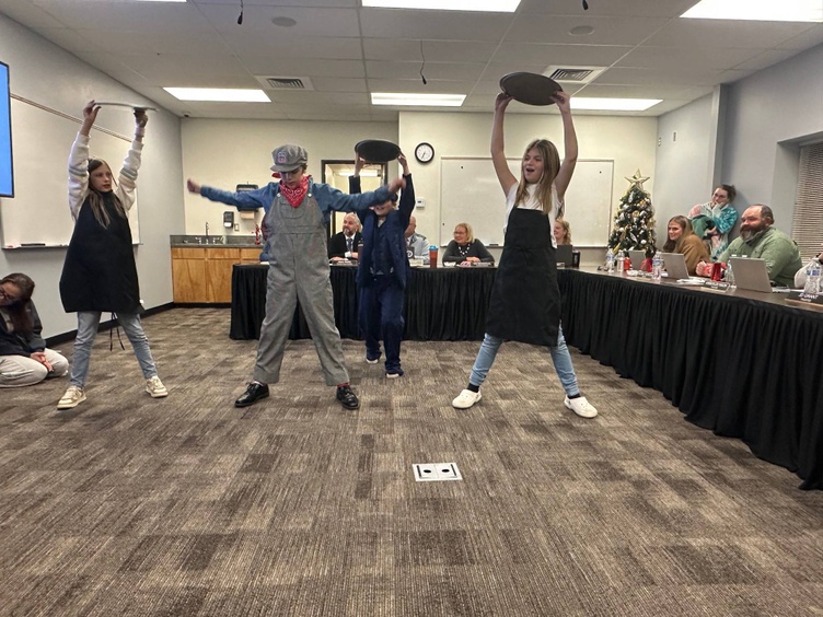 Four students, including one dressed as a train engineer, perform at a board meeting. The students are wearing aprons and holding trays raised above their heads in a choreographed motion. A Christmas tree is visible in the background, along with board members seated at a long table, smiling and observing the performance.