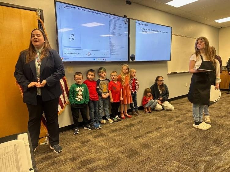 A group of young elementary students from Crimson Point Elementary stand in a line in front of a presentation screen at a board meeting, smiling. A teacher stands to the left, and a student wearing an apron stands to the right, holding a tray. The screen displays part of the meeting agenda, and some adults are seated in the background.