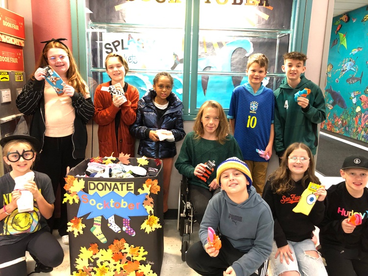 A group of elementary school students and an adult teacher pose with colorful socks they’ve collected for "Socktober," a donation drive. The students stand around a decorated donation bin filled with socks, featuring fall leaves and a banner that says "DONATE Socktober." The group smiles proudly in front of a bulletin board and a vibrant mural with underwater themes in the background.