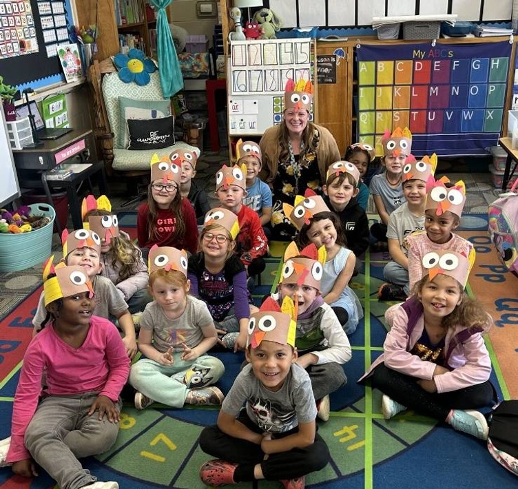 Kindergarten students sit on a floor rug with their teacher, all smiling and wearing handmade construction paper turkey hats