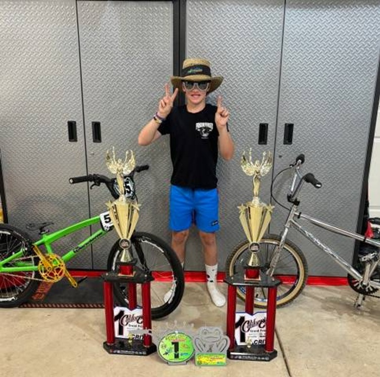 Student poses for the camera holding up 3 fingers, standing with 4 first-place trophies